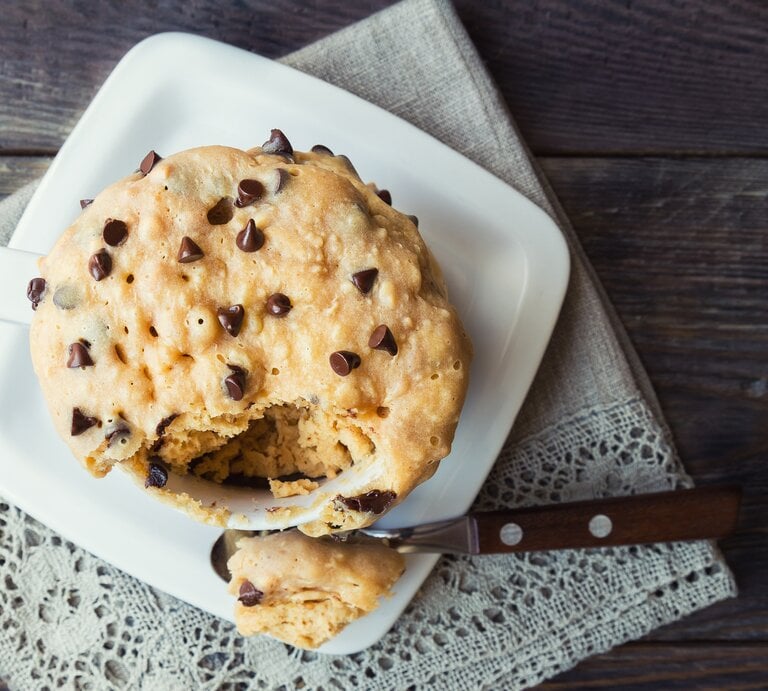 Eine Tasse mit gebackenen Haferflocken mit Banane und Schoko-Chips auf einem weißen Tassenuntersetzer neben einem Löffel auf einem verzierten Tuch vor einem Holzhintergrund, von oben.  | © Getty Images/ Anaiz777