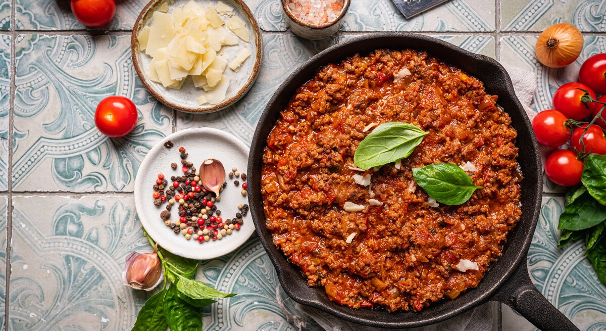 Gusseiserne Pfanne  mit Bolognese-Soße auf gekachelter Oberfläche. Umgeben ist der Topf von typischen Zutaten einer klassischen Bolognese. 