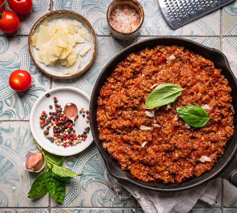Gusseiserne Pfanne  mit Bolognese-Soße auf gekachelter Oberfläche. Umgeben ist der Topf von typischen Zutaten einer klassischen Bolognese.  | © AdobeStock/petrrgoskov