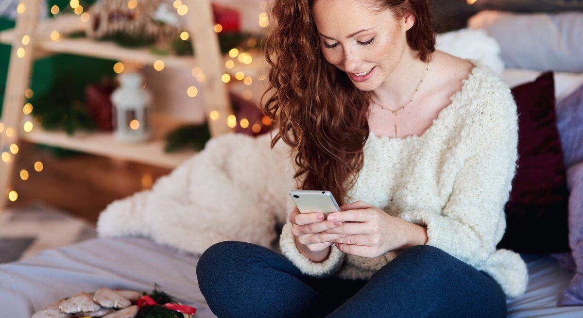 Junge Frau sitzt auf dem Sofa und verschickt Advent Sprüche an ihre Freunde und die Familie. Ihre Wohnung ist weihnachtlich dekoriert. 