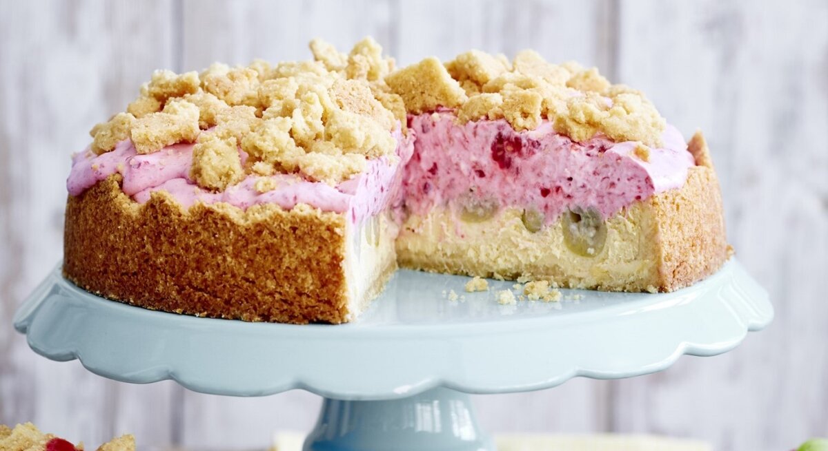 Auf einer Etagerie steht eine Stachelbeer-Quark-Streuseltorte. Sie ist mit einer rosa Creme gefüllt und angeschnitten. 