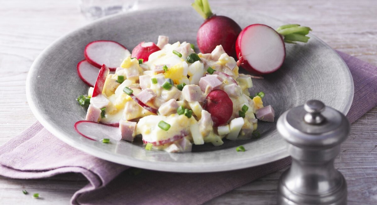 Eiersalat mit Pute und Radieschen in hellgrauer Schale auf lila Tuch und hellem Untergrund. Im Hintergrund ein Glas Wasser. Frontalaufnahme.