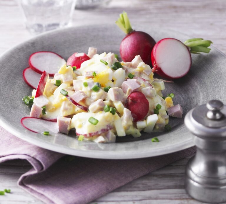 Eiersalat mit Pute und Radieschen in hellgrauer Schale auf lila Tuch und hellem Untergrund. Im Hintergrund ein Glas Wasser. Frontalaufnahme. | © StockFood / Gräfe & Unzer Verlag / Kramp + Gölling