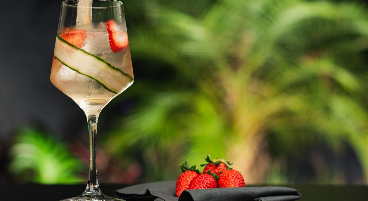 Ein Glas mit dem Lillet Vive auf dunklem Untergrund und vor vielen unscharfen Pflanzen im Hintergrund.
