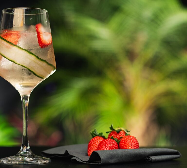 Ein Glas mit dem Lillet Vive auf dunklem Untergrund und vor vielen unscharfen Pflanzen im Hintergrund. | © Shutterstock/ Fernanda Flugel 
