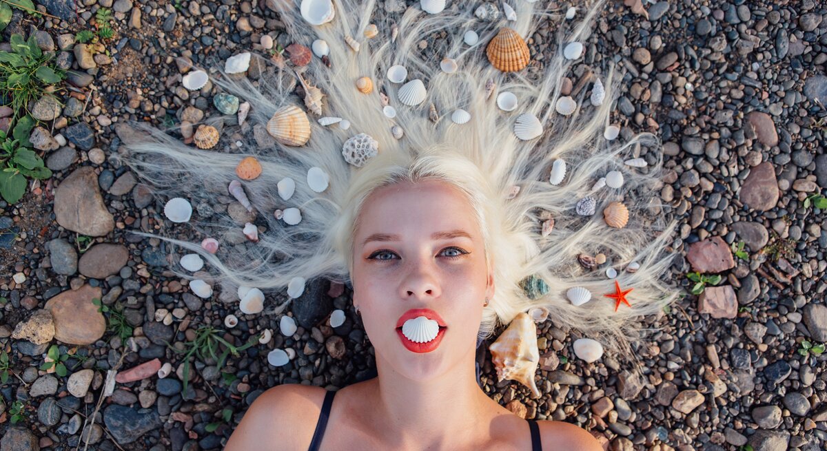Eine blonde Frau liegt mit dem Rücken auf einem Kieselstrand, umgeben von verschiedenen Meeresmuscheln, die in ihrem ausgebreiteten Haar arrangiert sind, während sie eine Muschelschale zwischen ihren Lippen hält, die wie ein Mund aussieht.