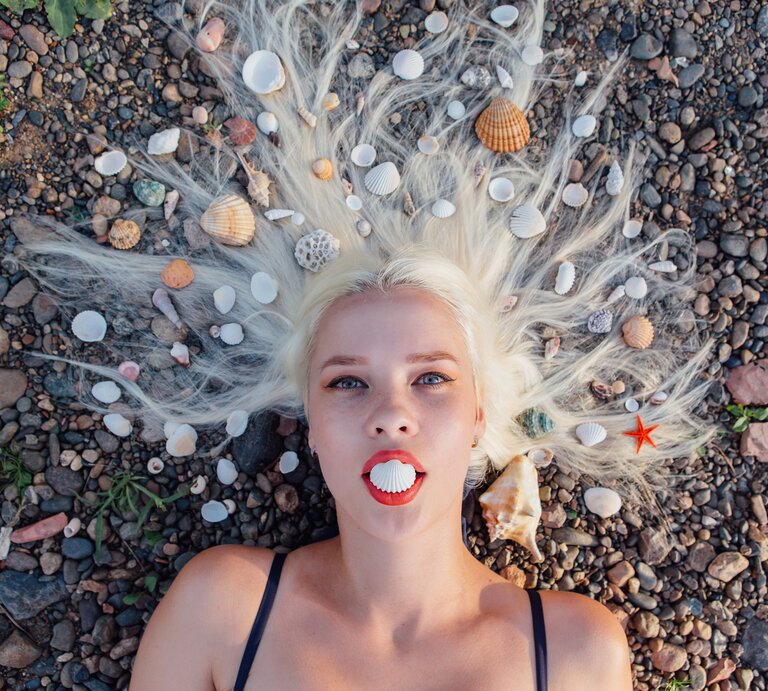 Eine blonde Frau liegt mit dem Rücken auf einem Kieselstrand, umgeben von verschiedenen Meeresmuscheln, die in ihrem ausgebreiteten Haar arrangiert sind, während sie eine Muschelschale zwischen ihren Lippen hält, die wie ein Mund aussieht. | © IMAGO / agefotostock