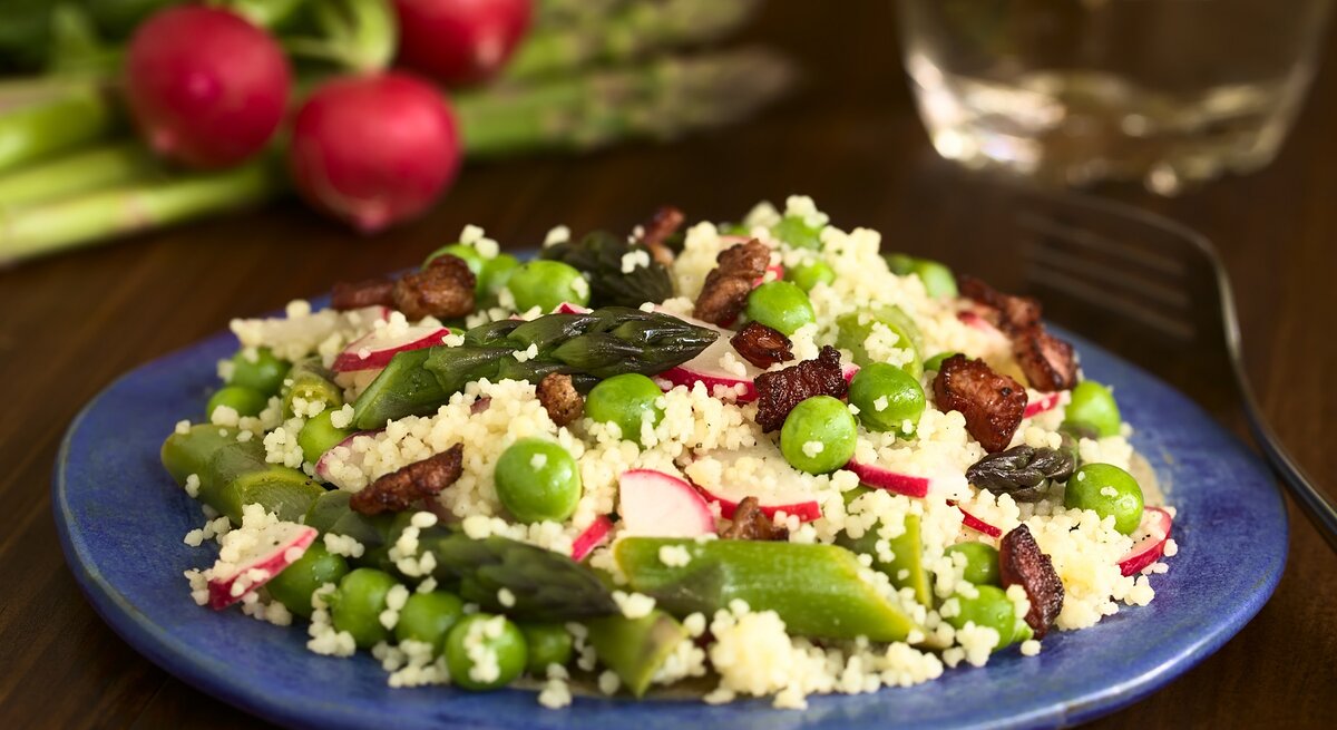 Eine Schale Spargel-Couscous-Salat mit Radieschen und Erbsen und Rosinen, drumherum die frischen Zutaten.