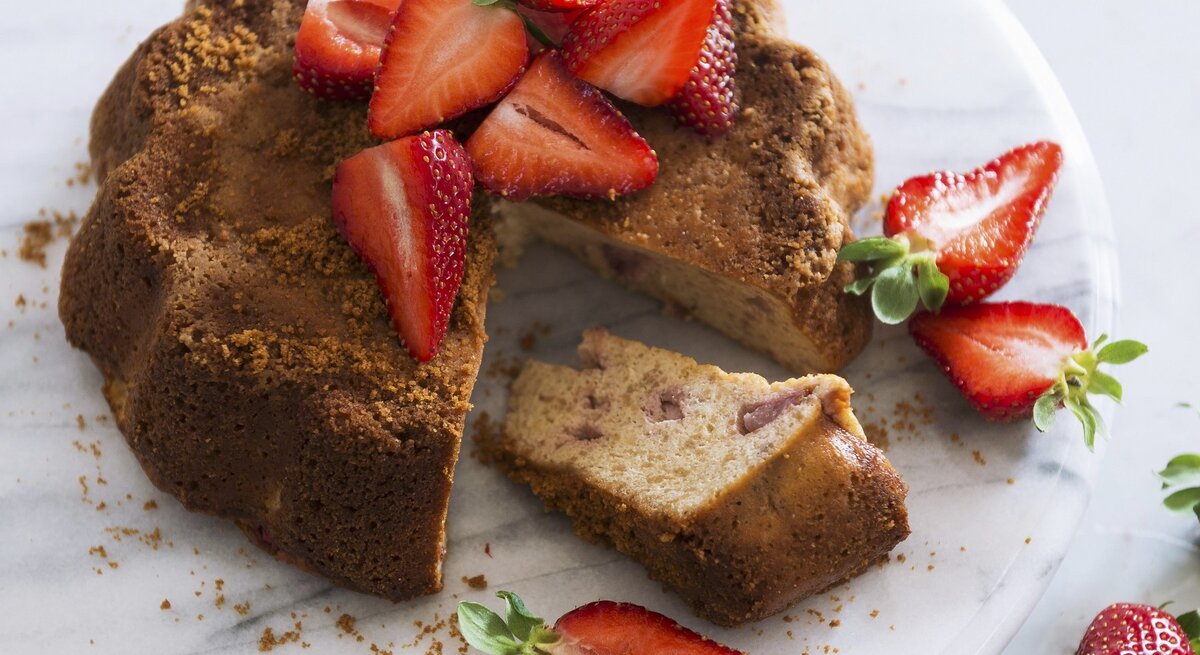Ein angeschnittener Sauerrahmkuchen mit Erdbeeren auf einem hellen Marmorteller und weißem Untergrund.