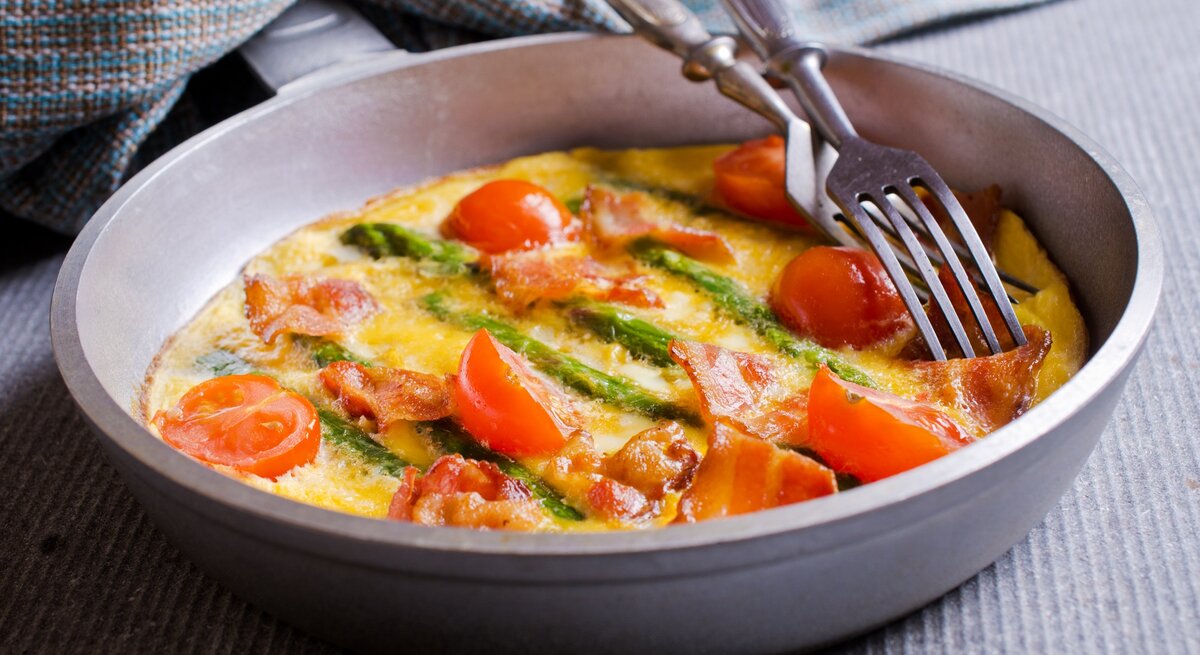 Eine Frittata mit grünem Spargel und Speck sowie Tomaten in einer Pfanne, darauf liegen zwei Gabeln, daneben ein Geschirrtuch.
