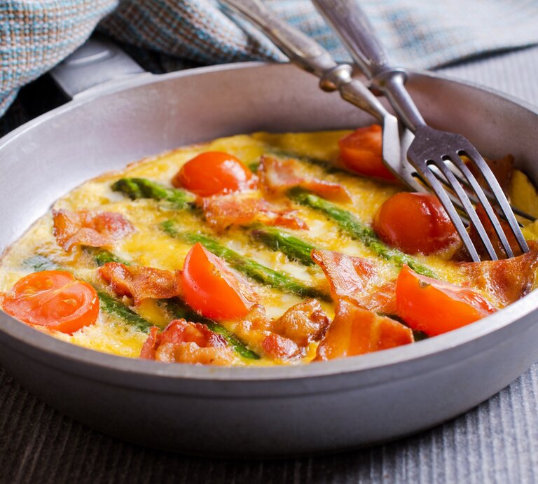 Eine Frittata mit grünem Spargel und Speck sowie Tomaten in einer Pfanne, darauf liegen zwei Gabeln, daneben ein Geschirrtuch. | © Adobe Stock/ Veronika Idiyat