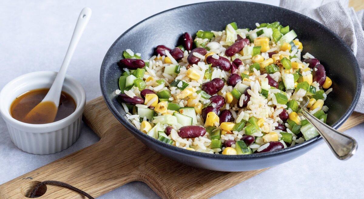 Reissalat mit Mais und Kidneybohnen in einer schwarzen Schüssel. Daneben ein weißes Schälchen mit einem Dressing. 