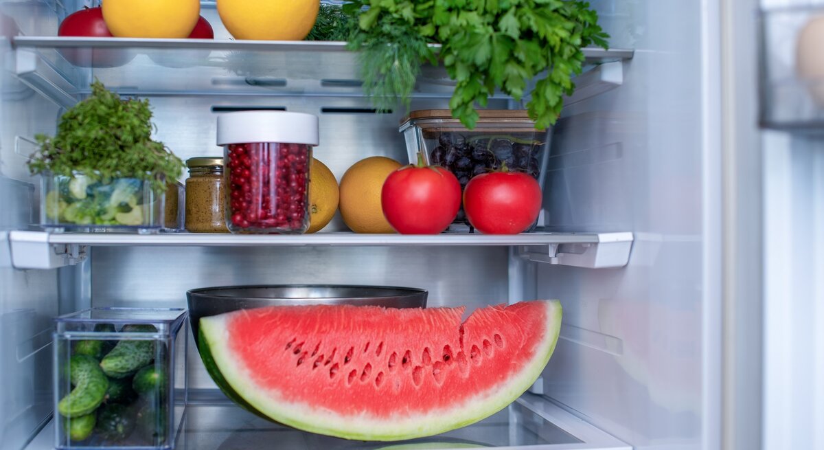 Ein geöffneter Kühlschrank in dem eine angeschnittene Wassermelone, Petersilie, Tomaten, Orangen und andere Lebensmittel gelagert werden.