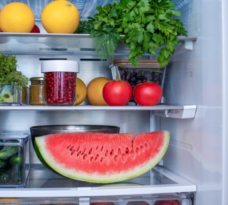 Ein geöffneter Kühlschrank in dem eine angeschnittene Wassermelone, Petersilie, Tomaten, Orangen und andere Lebensmittel gelagert werden. | © Shutterstock / Zakharova_Elena