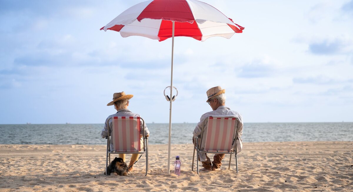 Zwei ältere Personen sitzen entspannt auf Strandstühlen unter einem rot-weißen Sonnenschirm am Strand. Sie tragen Strohhüte und sind dem Meer zugewandt, das sich im Hintergrund erstreckt. Daneben sitzt ein kleiner Hund im Sand.