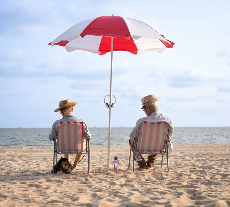 Zwei ältere Personen sitzen entspannt auf Strandstühlen unter einem rot-weißen Sonnenschirm am Strand. Sie tragen Strohhüte und sind dem Meer zugewandt, das sich im Hintergrund erstreckt. Daneben sitzt ein kleiner Hund im Sand. | © Getty Images / Patchareeporn Sakoolchai