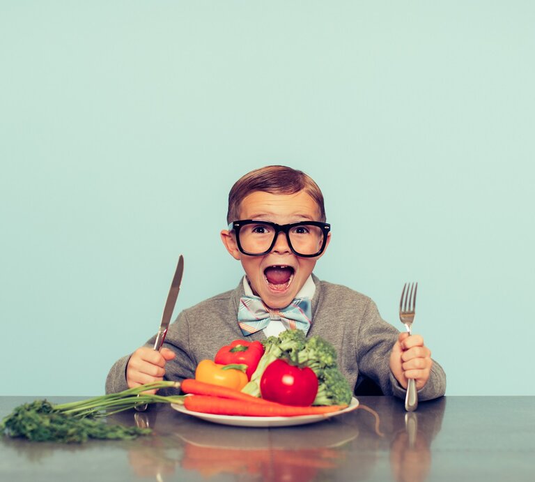 Vegane Ernährung bei Kindern: Ein Junge mit großer Brille und Fliege sitzt mit Besteck vor einem Teller mit Obst und Gemüse. | © GettyImages/ RichVintage