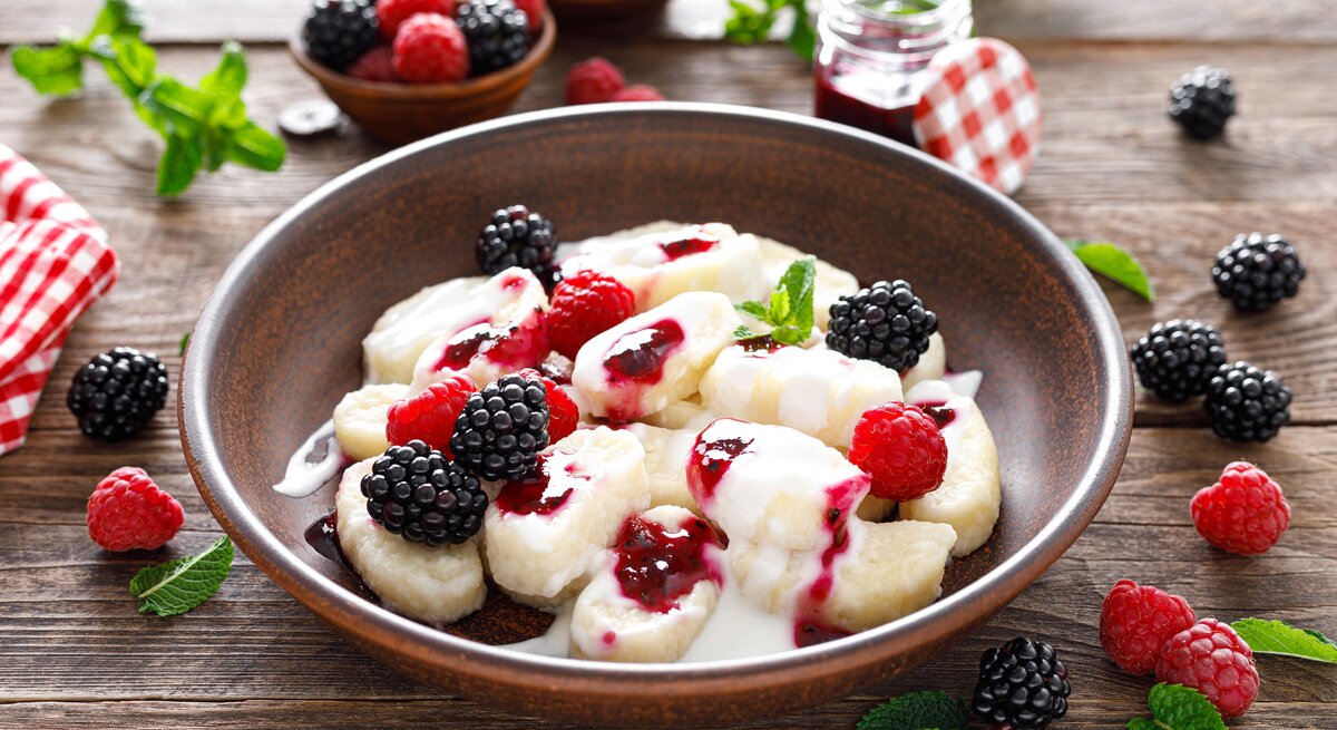 Brauner Teller gefüllt mit süßen Gnocchi, Sahne und Beeren. Darum herum liegen auf einer Holzplatte Beeren und Minze. 