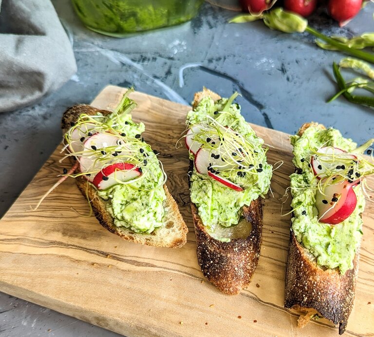 Ein kleines Holzbrett mit drei Scheiben Brot und dem Bärlauch-Aufstrich. Im Hintergrund wilder Bärlauch mit Radieschen und einem grauen Tuch. | © EAT CLUB/ Felix Scheel