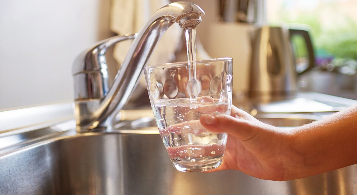 Das Bild zeigt eine Person, die ein Glas mit klarem Wasser aus einem modernen, metallischen Wasserhahn in einer Küchenspüle füllt. Das Tageslicht fällt durch ein Fenster ein und beleuchtet die Szene, was auf eine häusliche und gesunde Umgebung hinweist. Das Wasser sieht sauber aus und die Szene kommuniziert die Zugänglichkeit von sauberem Trinkwasser, was für eine gesunde Lebensweise unerlässlich ist.
