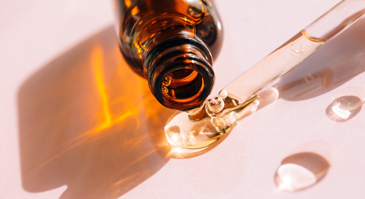Das Bild zeigt eine Nahaufnahme einer Pipette, die eine klare Flüssigkeit tropft, und eine braune Glasflasche, aus der die Flüssigkeit kommt. Die Flasche und die Pipette werfen aufgrund des durchsichtigen Öls einen orangefarbenen Schatten auf den hellen Untergrund. Dieses Setup ist typisch für ästhetische und hautpflegerelevante Produkte wie Seren oder Öle, die für ihre feuchtigkeitsspendenden oder anti-aging Eigenschaften vermarktet werden. Squalan ist eine Substanz, die bekannt dafür ist, die Haut zu hydratisieren und wird oft in hautpflege Produkten verwendet.