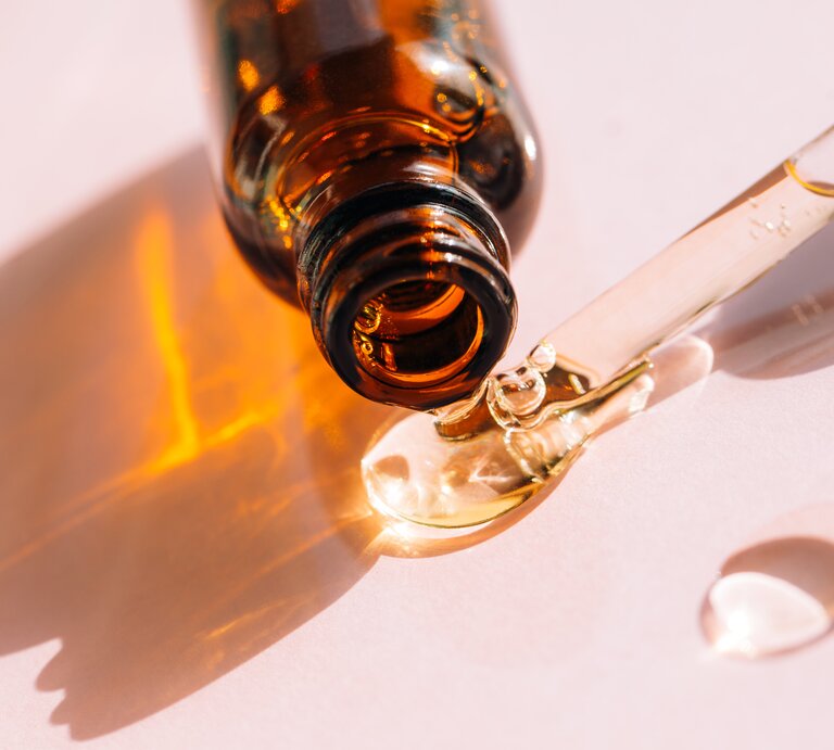 Das Bild zeigt eine Nahaufnahme einer Pipette, die eine klare Flüssigkeit tropft, und eine braune Glasflasche, aus der die Flüssigkeit kommt. Die Flasche und die Pipette werfen aufgrund des durchsichtigen Öls einen orangefarbenen Schatten auf den hellen Untergrund. Dieses Setup ist typisch für ästhetische und hautpflegerelevante Produkte wie Seren oder Öle, die für ihre feuchtigkeitsspendenden oder anti-aging Eigenschaften vermarktet werden. Squalan ist eine Substanz, die bekannt dafür ist, die Haut zu hydratisieren und wird oft in hautpflege Produkten verwendet. | © Getty Images/Anna Efetova