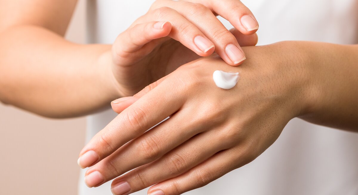 Das Bild zeigt die Hände einer Person, die eine Portion Feuchtigkeitscreme auf dem Handrücken hat. Die Hände haben gepflegte Nägel und die Haut erscheint gesund. Die Creme ist bereit, in die Haut einmassiert zu werden, was auf eine Hautpflegeroutine hinweist. Der Hintergrund ist neutral und lässt den Fokus auf die Handpflege.