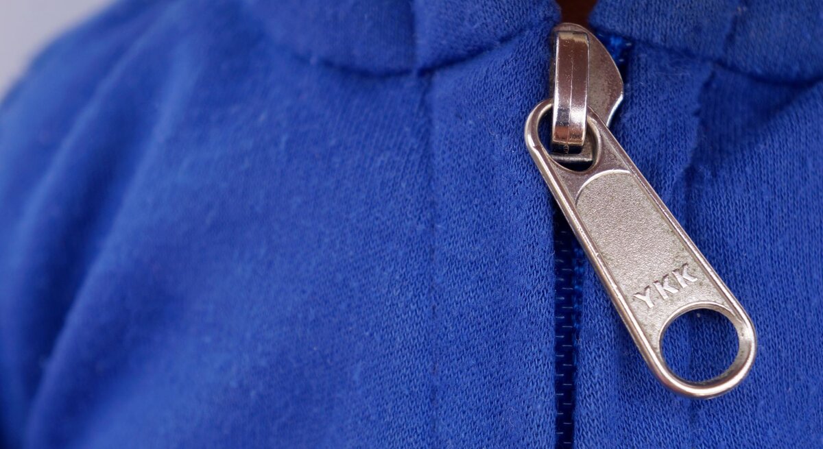 Nahaufnahme eines Reißverschlusses an einem blauen Kleidungsstück. Auf dem silbernen Zipper ist der Schriftzug "YKK" eingraviert.