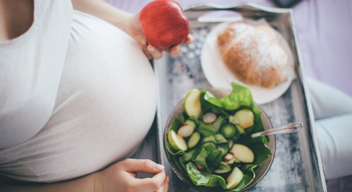 Das Bild zeigt eine schwangere Frau, die auf einem Bett sitzt. Ihr großer Babybauch ist unter einem engen, weißen Oberteil zu erkennen. Sie hält einen roten Apfel in ihrer rechten Hand und hat ein Holztablett auf ihren Knien, auf dem eine Schüssel mit grünem Salat und ein Teller mit einem Croissant liegen. Die Szene strahlt eine ruhige, häusliche Atmosphäre aus und betont die Bedeutung einer gesunden Ernährung während der Schwangerschaft.