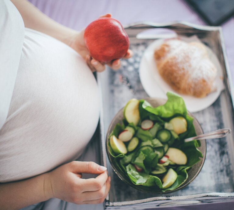 Das Bild zeigt eine schwangere Frau, die auf einem Bett sitzt. Ihr großer Babybauch ist unter einem engen, weißen Oberteil zu erkennen. Sie hält einen roten Apfel in ihrer rechten Hand und hat ein Holztablett auf ihren Knien, auf dem eine Schüssel mit grünem Salat und ein Teller mit einem Croissant liegen. Die Szene strahlt eine ruhige, häusliche Atmosphäre aus und betont die Bedeutung einer gesunden Ernährung während der Schwangerschaft. | © iStock/Pekic