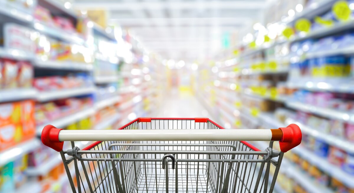 Ein leerer Einkaufswagen in einem Supermarktgang. Die Produkte sind unscharf zu erkennen.