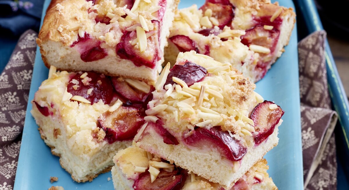 Pflaumen-Butterkuchen vom Blech, geschnitten und aufeinander auf einem blauen Teller gestapelt serviert. Die einzelnen Kuchenstücke mit Pflaumen und Mandelsplittern garniert. 
