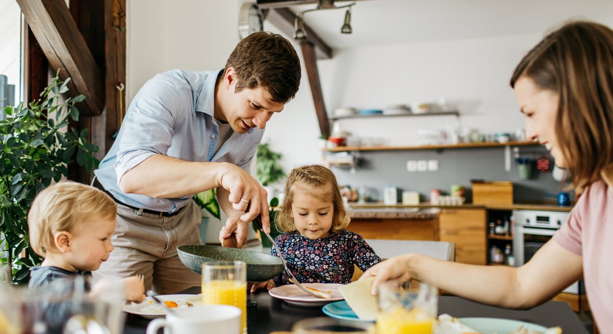 Eine glückliche Familie beim Frühstück: Ein Mann in einem hellblauen Hemd beugt sich über, um seiner kleinen Tochter zu helfen, die konzentriert auf ihren Teller blickt. Ein weiteres Kleinkind sitzt daneben und ist auf sein Essen fokussiert. Eine Frau sitzt gegenüber, reicht etwas hinüber und lächelt dabei. Der Tisch ist mit einem gesunden Frühstück mit Orangensaft, Spiegeleiern und Brötchen gedeckt. Sie befinden sich in einer hellen Küche mit moderner Einrichtung und Pflanzen, die für eine wohnliche Atmosphäre sorgen.
