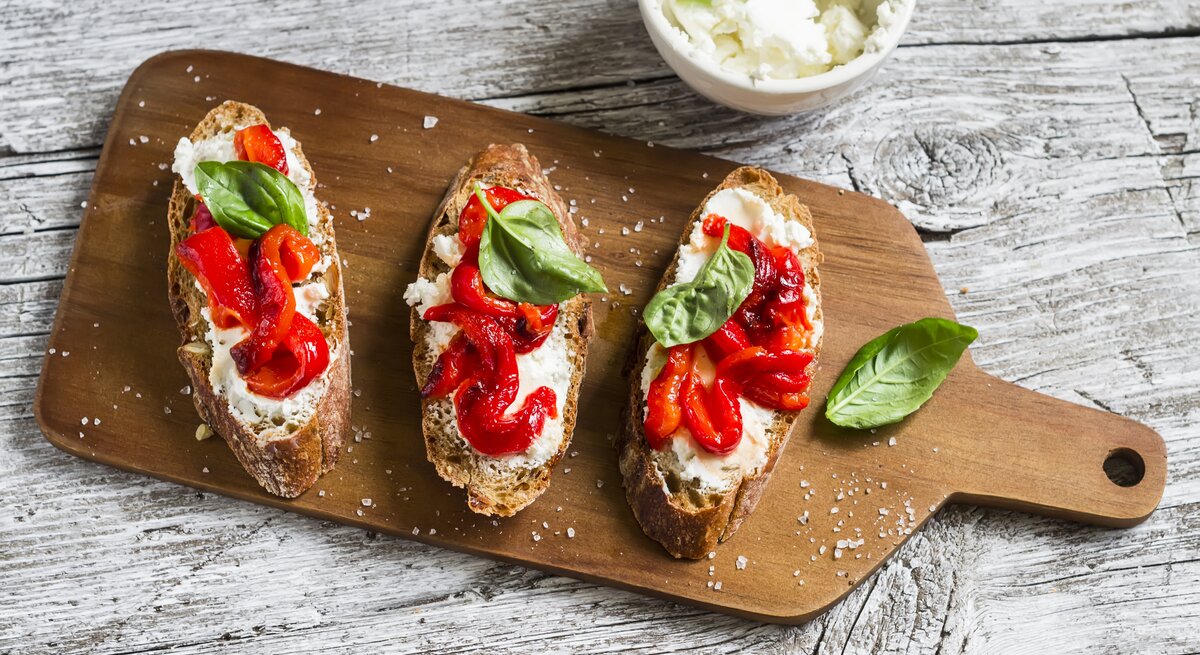 Auf einem Holzbrettchen lieben drei Scheiben Knoblauch-Brot mit Paprika und Ricotta. Auf jedem Brot ist ein Blättchen Basilikum.