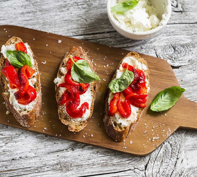Auf einem Holzbrettchen lieben drei Scheiben Knoblauch-Brot mit Paprika und Ricotta. Auf jedem Brot ist ein Blättchen Basilikum. | © Shutterstock / Kiian Oksana