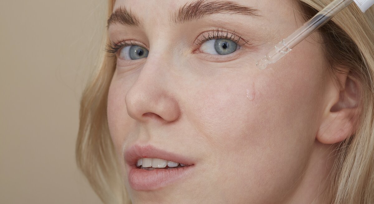 Ein Bild einer Frau mit heller Haut und blonden Haaren, die ein Serum mit einer Pipette auf ihre Wange aufträgt. Die Flüssigkeit ist gerade dabei, in kleinen Tropfen entlang ihrer Wange herunterzulaufen. Sie hat hellblaue Augen und trägt ein minimales Make-up. Ihre Mimik wirkt entspannt und der Gesichtsausdruck ist ruhig. Der Hintergrund ist neutral und in einem warmen Beigeton gehalten, was die klare Fokussierung auf die Anwendung des Serums unterstützt.