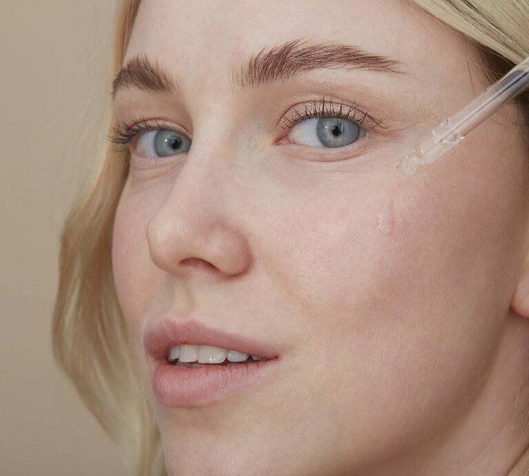 Ein Bild einer Frau mit heller Haut und blonden Haaren, die ein Serum mit einer Pipette auf ihre Wange aufträgt. Die Flüssigkeit ist gerade dabei, in kleinen Tropfen entlang ihrer Wange herunterzulaufen. Sie hat hellblaue Augen und trägt ein minimales Make-up. Ihre Mimik wirkt entspannt und der Gesichtsausdruck ist ruhig. Der Hintergrund ist neutral und in einem warmen Beigeton gehalten, was die klare Fokussierung auf die Anwendung des Serums unterstützt. | © Getty Images/Westend61