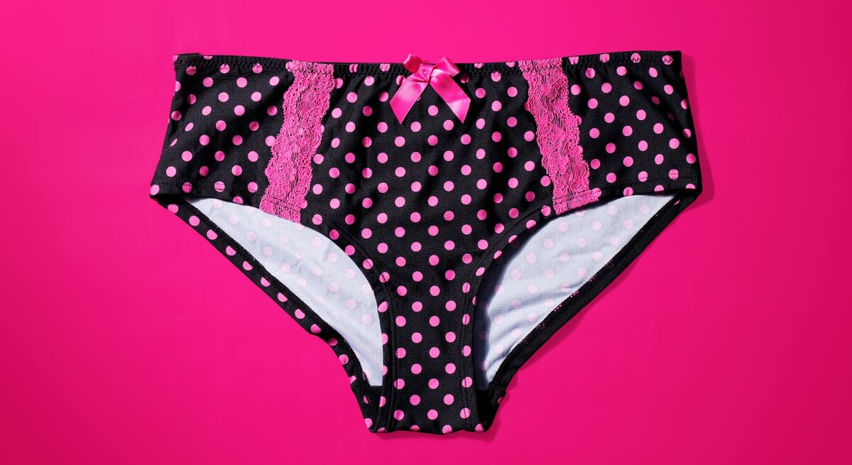 Schwarzer Slip mit pinkfarbenen Punkten und Spitzenverzierungen auf einem knallpinken Hintergrund. Der Slip ist zudem mit einer kleinen pinkfarbenen Schleife an der Vorderseite dekoriert.