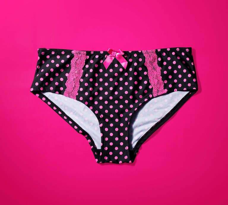 Schwarzer Slip mit pinkfarbenen Punkten und Spitzenverzierungen auf einem knallpinken Hintergrund. Der Slip ist zudem mit einer kleinen pinkfarbenen Schleife an der Vorderseite dekoriert. | © Getty Images /	stocksnapper