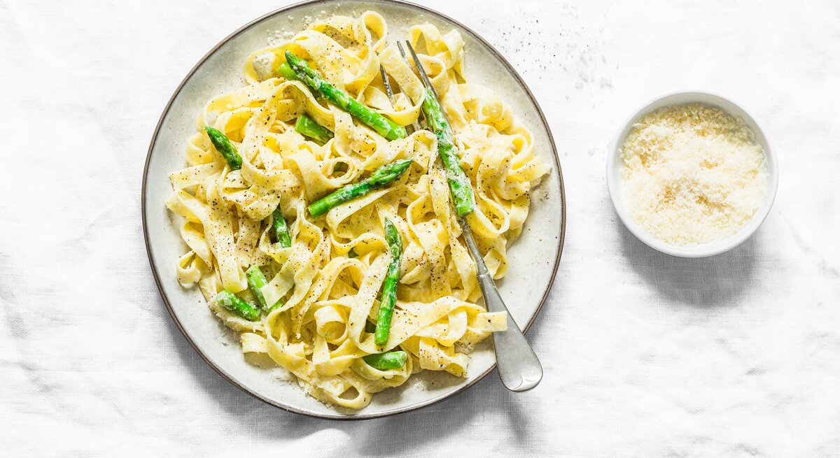 Ein Teller Frischkäse-Pasta mit grünem Spargel in der Draufsicht, daneben eine Gabel und eine Schale Parmesan.