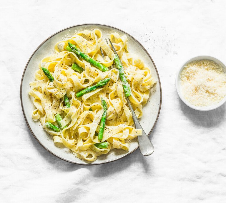 Ein Teller Frischkäse-Pasta mit grünem Spargel in der Draufsicht, daneben eine Gabel und eine Schale Parmesan. | © Adobe Stock/ okkijan2010