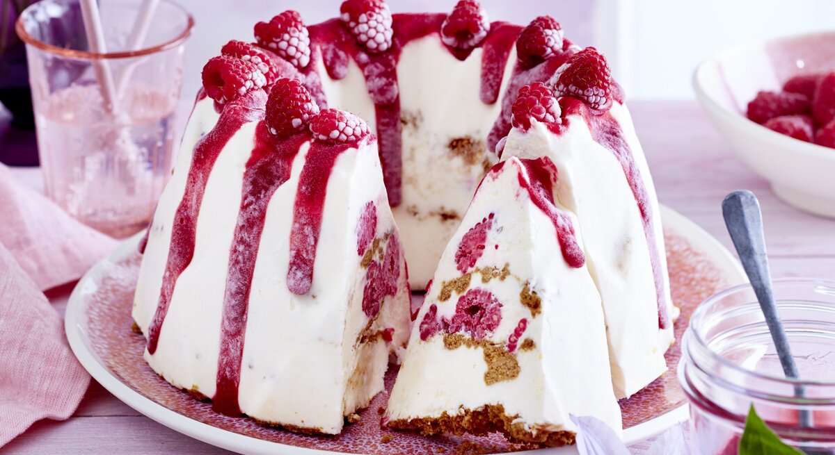 Cheesecake-Eis-Gugelhupf, mit einem Boden aus knusprigem Keks und einem Topping aus Himbeersauce und frischen Himbeeren, serviert auf einem hellen Tisch auf weißem Teller. 
