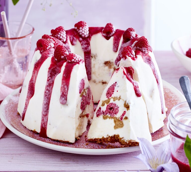 Cheesecake-Eis-Gugelhupf, mit einem Boden aus knusprigem Keks und einem Topping aus Himbeersauce und frischen Himbeeren, serviert auf einem hellen Tisch auf weißem Teller.  | © EAT CLUB / Isabell Triemer
