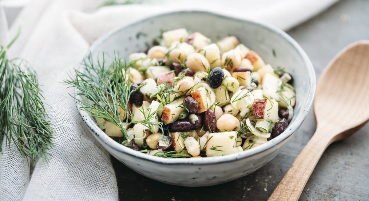 Eine Schüssel Apfel-Bohnensalat mit Kichererbsen und Dill, daneben ein Holzlöffel.