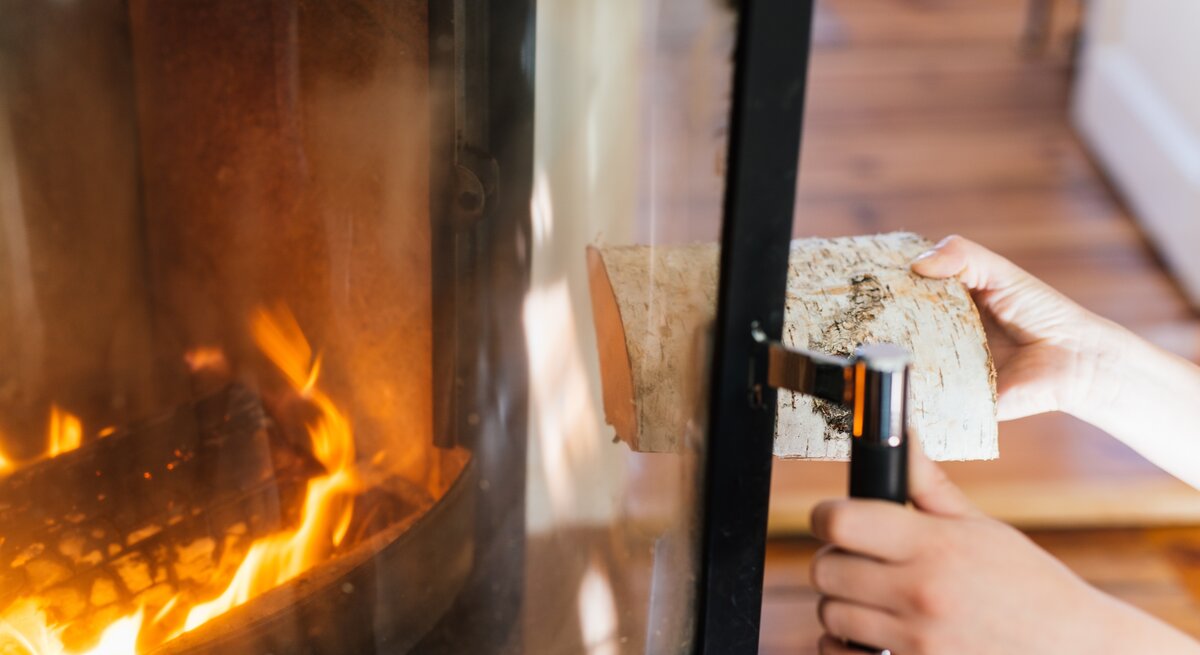 Person öffnet Kaminscheibe und wirft Holz in das Feuer des Kamins.