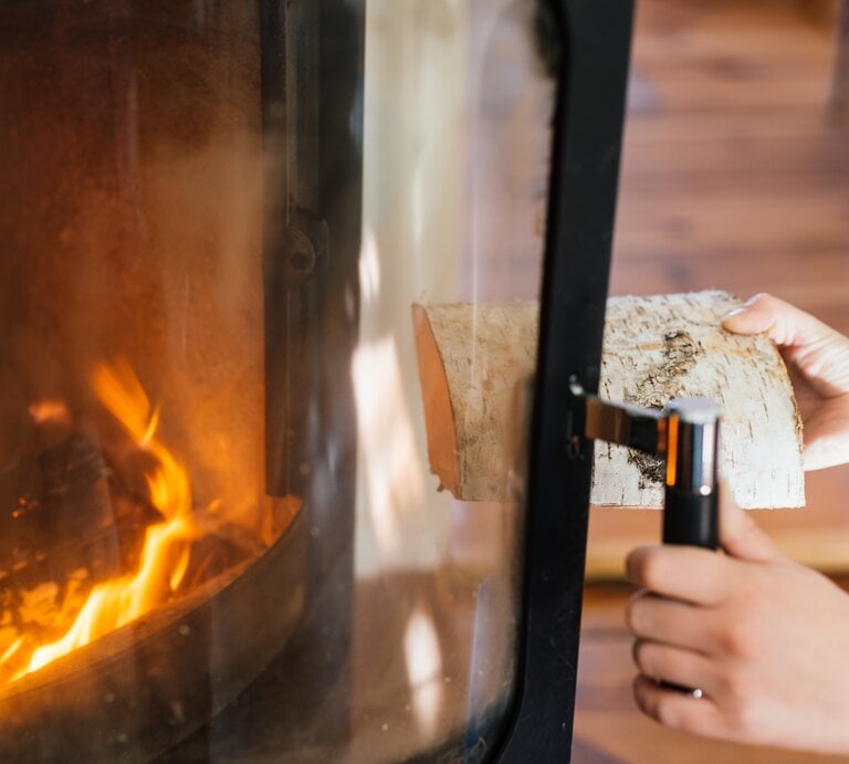Person öffnet Kaminscheibe und wirft Holz in das Feuer des Kamins. | © Getty Images/Guido Mieth