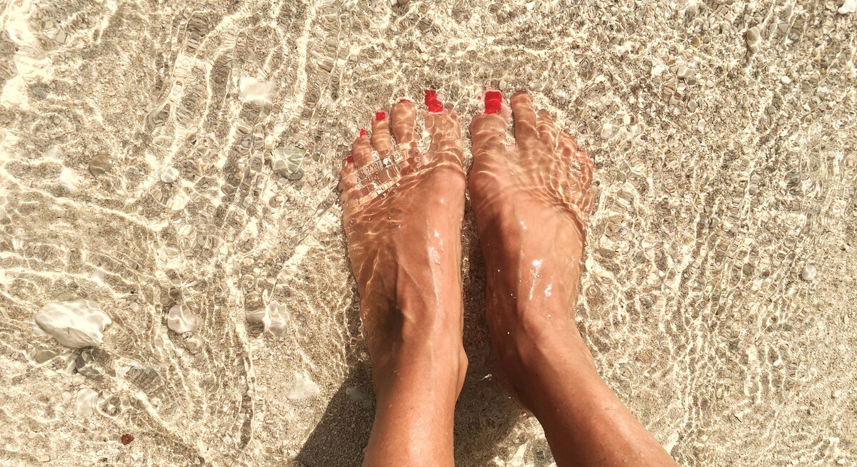 Füße einer Person, die im klaren, flachen Meerwasser am Strand stehen. Die Zehen sind mit rotem Nagellack lackiert. Man sieht, wie das Sonnenlicht das Wasser auf dem sandigen Untergrund funkeln lässt.