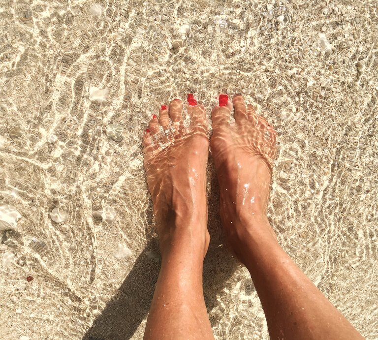 Füße einer Person, die im klaren, flachen Meerwasser am Strand stehen. Die Zehen sind mit rotem Nagellack lackiert. Man sieht, wie das Sonnenlicht das Wasser auf dem sandigen Untergrund funkeln lässt. | © iStock/irenetinta
