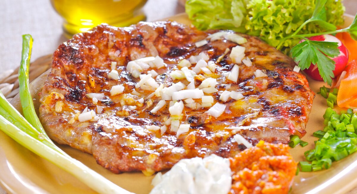 Pljeskavica kroatischer Hamburger mit Schafskäsefüllung und Beilgane und Holzplatte und hellem Untergrund. Daneben Olivenöl. Draufsicht.
