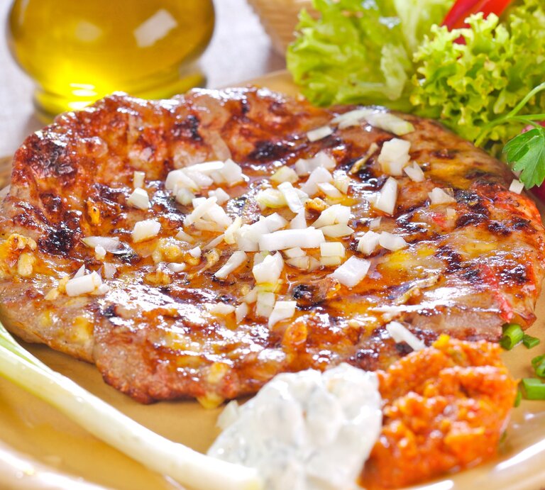 Pljeskavica kroatischer Hamburger mit Schafskäsefüllung und Beilgane und Holzplatte und hellem Untergrund. Daneben Olivenöl. Draufsicht. | © Getty Images / ShyMan